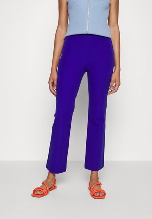 Vrouw die felblauwe broek met rechte pijpen draagt, een mouwloze lichtblauwe top en oranje sandalen met bandjes, poseert voor een effen achtergrond.