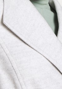 Primer plano de una solapa de abrigo de lana gris claro sobre un suéter de cuello alto de punto acanalado verde pálido.