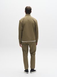 Khaki värvi jope püstkraega, sobitatud sama värvi pükstega. Mõlemal rõival on sile ja mati tekstuuriga pind. Välimuse täiendab mustad tossud.