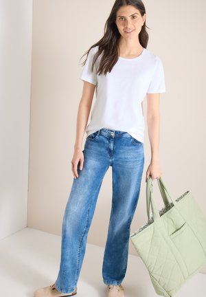 Femme portant un t-shirt blanc et un jean bleu tenant un grand sac fourre-tout matelassé vert, debout contre un mur beige.
