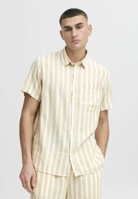 Camisa de manga corta abotonada a rayas beige y blancas. Fabricada en un tejido ligero, cuenta con un bolsillo en el pecho y un corte relajado.