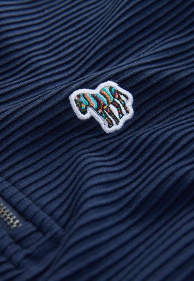 Tumši zils ribots audums, uz kura ir krāsains zebras izšuvuma logo ar baltu kontūru un rāvējslēdzēja detaļa apakšējā kreisajā stūrī.