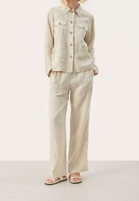 Tenue en lin beige clair comprenant une chemise boutonnée avec deux poches poitrine et un pantalon à jambes larges. Des sandales à semelle plate complètent le look.