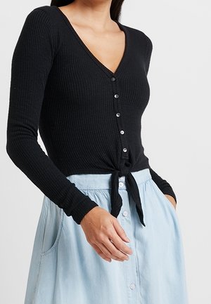 Czarna prążkowana bluzka z długim rękawem, dekoltem w szpic, zapięciem na guziki z przodu oraz detalem wiązania w talii, zestawiona z jasnoniebieską spódnicą zapinaną na guziki.