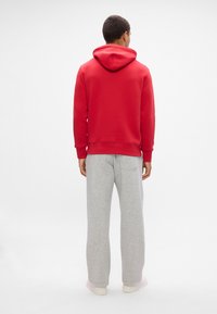 Homme de dos, portant un sweat à capuche rouge, un pantalon de survêtement gris clair et des baskets blanches, sur un fond blanc uni.