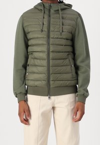 Grøn hættejakke med quiltet front, ribbede manchetter og justerbare snore. Har sidelommer og lynlåslukning.