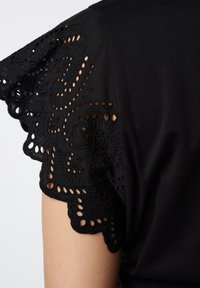 Top noir avec des manches en dentelle bordées de motifs découpés complexes et de bords festonnés, fabriqué en tissu doux et respirant.
