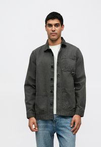 Γκρι βαμβακερό μπουφάν με γιακά, κλείσιμο με κουμπιά, τσέπη στο στήθος και δύο μπροστινές τσέπες, φορεμένο πάνω από άσπρο πουκάμισο και μπλε τζιν.