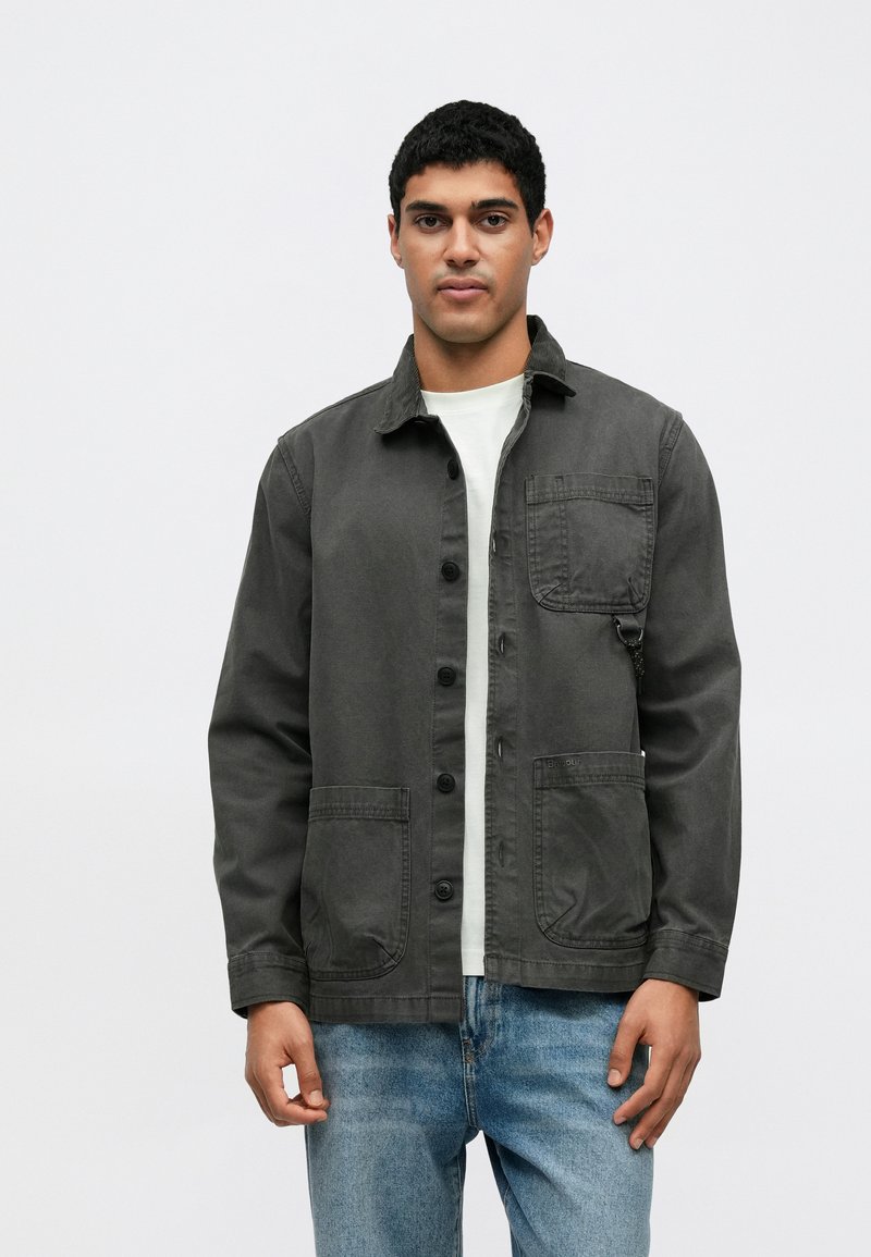 Γκρι βαμβακερό μπουφάν με γιακά, κλείσιμο με κουμπιά, τσέπη στο στήθος και δύο μπροστινές τσέπες, φορεμένο πάνω από άσπρο πουκάμισο και μπλε τζιν.