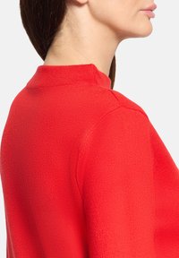 Kobieta w czerwonym dzianinowym swetrze widziana z boku i z tyłu, skupiając się na okolicy ramienia i szyi, z brązowymi włosami związanymi do tyłu.