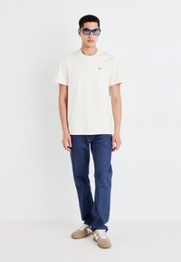 Kremfarget t-skjorte med korte ermer og en liten logo, paret med mørkeblå denimjeans og beige sneakers med hvite snørebånd. Tilbehøret inkluderer solbriller.