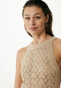 Beige stickad topp med ett sicksackmönster, hög krage och öppna sidor. Tyget har en strukturerad, lätt och luftig känsla.