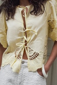 Rumena vezenina bluzica z širokimi rokavi, ki ima čipkaste izreze in zavezovanje na izrezu vratu. V kombinaciji z belo kratko hlače z teksturo.