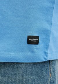 Světle modré bavlněné tričko s černou etiketou s nápisem "JACK & JONES EST. 1990" poblíž lemu, v kombinaci se světle modrými džíny.
