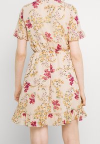 Kukallinen mekko vaaleassa beigessä, jossa on vaaleanpunaisia ja keltaisia kukkia. Siinä on lyhyet röyhelösateet, joustovyötärö ja röyhelöinen helma.