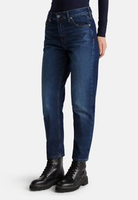 Jeans en denim bleu foncé avec une coupe droite, des détails de couture visibles et des poches avant. Associés à des bottes noires à lacets.