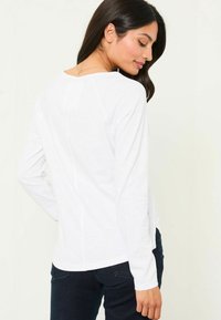 Weißes Langarmoberteil mit rundem Halsausschnitt und Rückennaht-Detail, kombiniert mit dunklen Jeans. Weicher Stoff, lässige Passform.