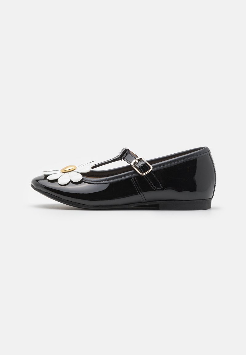 Chaussure Mary Jane noire en cuir verni avec boucle latérale et décoration marguerite blanche sur le bout arrondi, semelle plate et finition lisse.