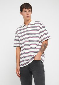 Polo de manga corta en crema con rayas horizontales en azul marino, burdeos y gris. Presenta un cuello clásico y tapeta de botones.