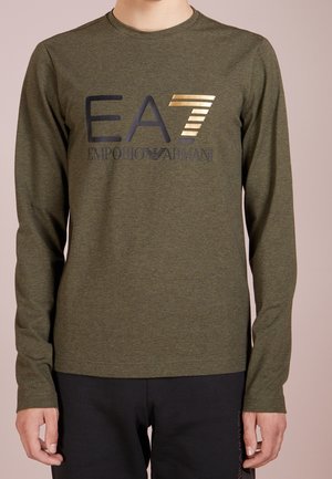 Langarmshirt in olijfgroen met het "EA7 Emporio Armani" logo in zwart en goud, gedragen door een staand persoon tegen een effen achtergrond.