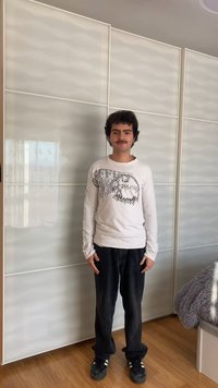 Junger Mann mit dunklem, lockigem Haar und Schnurrbart, trägt ein weißes langärmliges Hemd und schwarze Jeans, steht drinnen auf einem Holzboden.