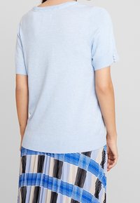 Maglione a maniche corte di colore blu chiaro in maglia con orlo a coste e accenti decorativi di perle sulle maniche. Sullo sfondo si intravede una gonna a fantasia.