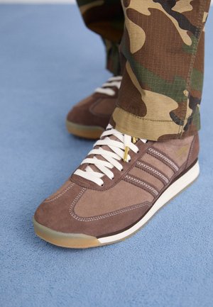 Zapatilla de ante marrón con cordones blancos usada con pantalones de camuflaje verdes y marrones sobre una superficie texturizada azul.