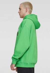 Sudadera verde con bolsillo canguro, capucha con cordón y puños acanalados. Presenta un detalle del logo en la parte frontal. Fabricada con un tejido suave y texturizado.