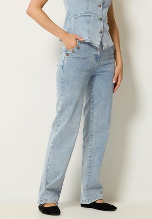 Kvinde iført lyseblå højtaljede jeans med knapdetaljer og matchende ærmeløs denimvest, kombineret med sorte flade sko.