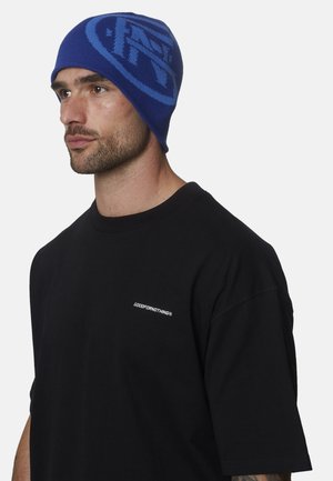 Hombre con un gorro de punto azul con un patrón y una camiseta negra con el logo de "GOODFORNOTHING" en el pecho, mirando hacia la izquierda.