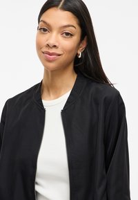 Νέα γυναίκα με μακριά σκούρα μαλλιά, φορώντας ασημένιες κρίκους, ένα λευκό μπλουζάκι και μια μαύρη φούτερ με φερμουάρ, που χαμογελά ελαφρώς σε λευκό φόντο.