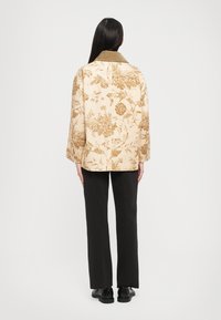 Ziedu raksta bēšs žakets ar olīvzaļo apkakli, brīvu struktūru un platiem piedurknēm, kombinēts ar melniem taisnām biksēm un apaviem.