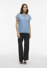 Blusa azzurra chiara con maniche corte e trasparenti, motivo a pois e dettaglio di un nodo sul collo, abbinata a pantaloni neri su misura e sandali neri.