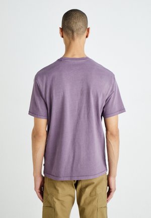 Homme aux cheveux courts portant un t-shirt violet uni à manches courtes et un pantalon kaki, debout de dos sur un fond uni.