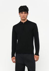 Zwarte polo met lange mouwen en een ritskraag. Gebreide textuur, slim fit ontwerp en geribde manchetten. Logopatch op de linkermouw.