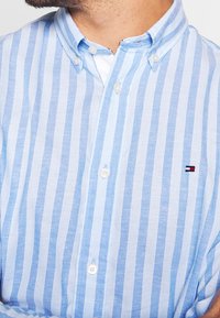 Light blue and white striped button-up shirt with a classic collar, featuring white buttons and a small embroidered logo on the chest.