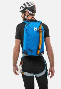 Blauer Rucksack mit strukturierten Oberflächen, Befestigungspunkte für das Geschirr und einer Eisaxt, getragen auf einem schwarzen Hemd mit Ausrüstungsringen und Riemen.