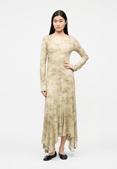 Γυναίκα που στέκεται με το ένα χέρι στη μέση, φορώντας μακρύ μπεζ φόρεμα με μοτίβο και ασύμμετρο τελείωμα, καθώς και μαύρα ίσια παπούτσια, μπροστά σε λευκό φόντο.