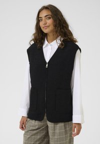 Crni vuneni prsluk s patentnim zatvaračem, s V-izrezom, dva prednja džepa i mekanom teksturom. Odjeven preko bijele košulje i uzorčenih hlača.