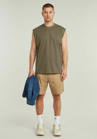 Maglietta senza maniche verde oliva, pantaloni corti beige con motivo, giacca di jeans appoggiata sul braccio, calzini bianchi e sneaker chunky.