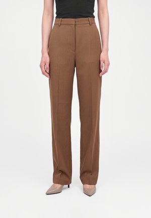 Femme portant un pantalon marron taille haute à jambes larges, un top noir sans manches et des talons beiges pointus, debout devant un fond uni.