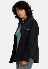 Donker corduroy overhemdjack met knopen, borstzak en klassieke kraag; gedragen over een lichtblauwe top en gecombineerd met denim.