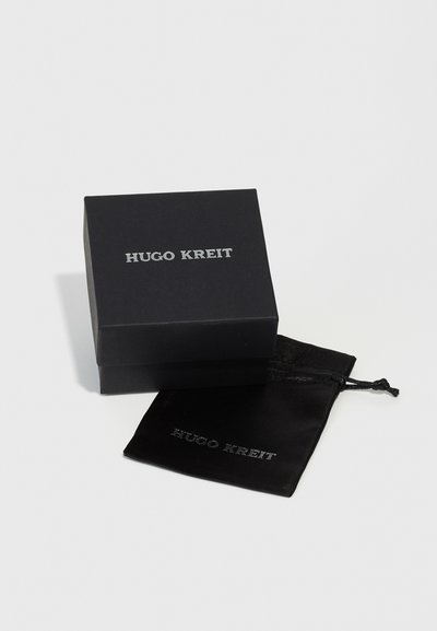 Boîte noire avec le texte argenté "Hugo Kreit" sur le dessus, accompagnée d'une pochette en velours noir portant le même texte. Texture lisse, forme carrée compacte.