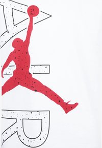 Biała koszulka z dużą czerwoną sylwetką skaczącej postaci sięgającej po koszykową piłkę, podkreślona czarnymi kropeczkami i konturami.