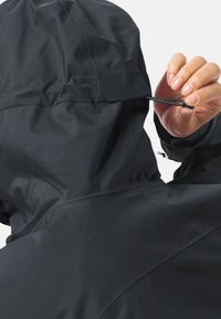 Zwarte waterdichte jas met een capuchon. De textuur is glad en de jas heeft een verstelbaar koord met een toggle aan de achterkant, wat het functionele ontwerp benadrukt.