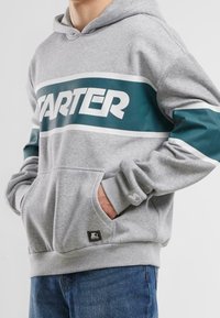 Sudadera gris con una banda verde azulado que presenta el logotipo "STARTER". Incluye un bolsillo delantero, puños acanalados y una pequeña etiqueta de marca en el dobladillo.