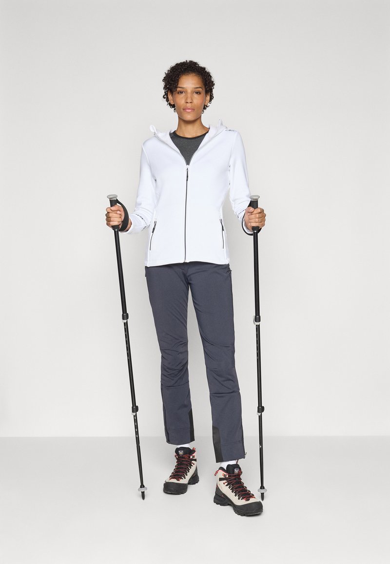 Giacca zip bianca, pantaloni grigio scuro, stivali neri con lacci rossi; tiene in mano i bastoncini da trekking e si trova davanti a uno sfondo semplice.