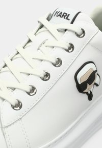 Vit sneaker med vita snören, metallögon, "KARL"-logotyp på tungan och ett tecknat ansikte med vitt hår och svarta solglasögon på sidan.