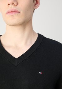 Pulover negru cu decolteu în V, cu guler rayat și un mic logo brodat în roșu, alb și albastru pe partea stângă a pieptului. Textură moale de tricot.