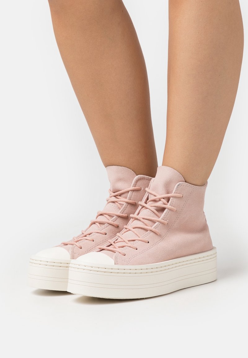 Jambes portant des baskets montantes en daim rose avec des semelles épaisses blanches et des lacets, debout sur une surface blanche.